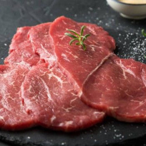 Raw marbled beef slices on a dark slate platter with rosemary sprigs and small bowls of seasoning in the background.