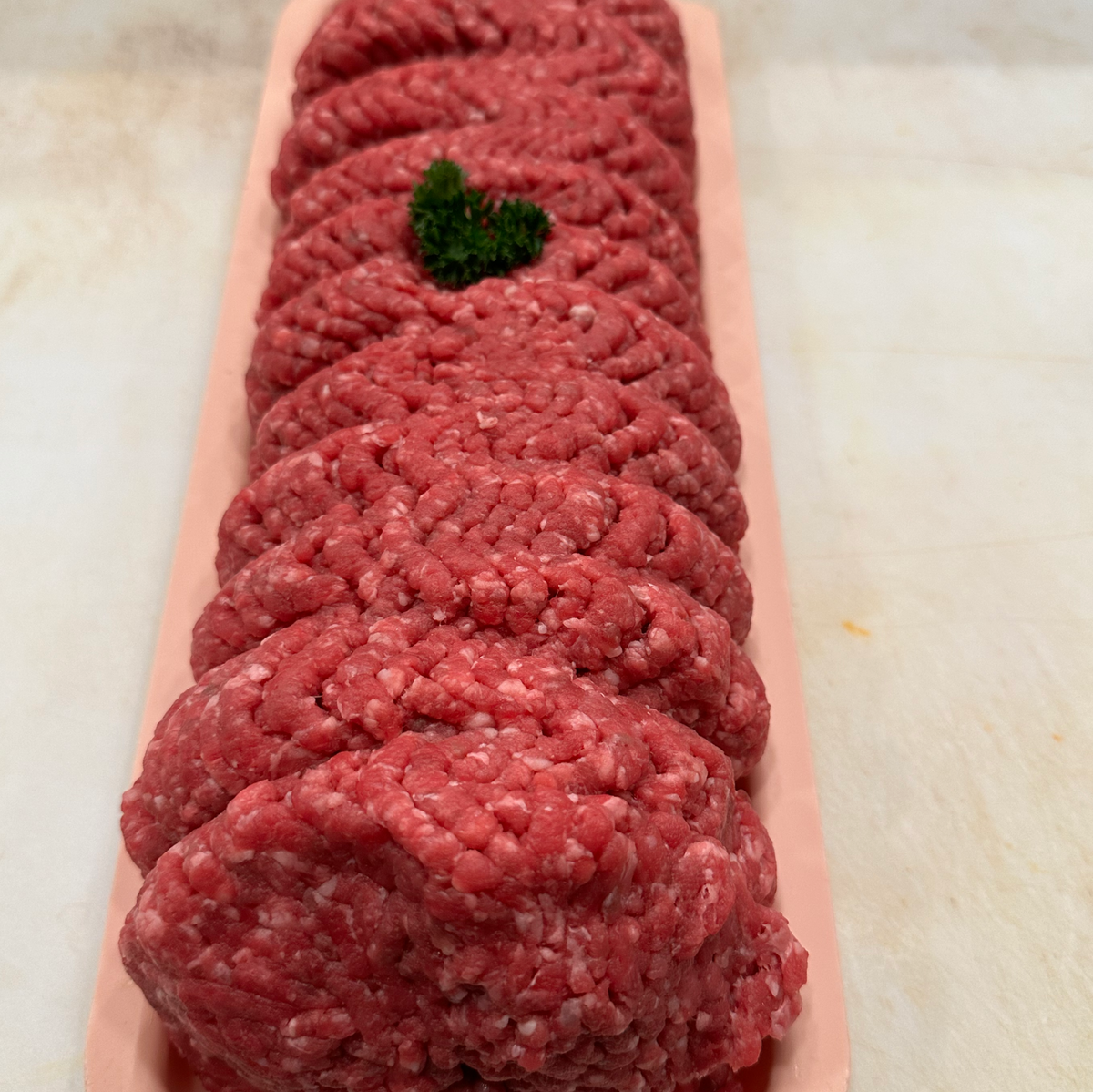 Raw ground meat patties arranged in a pink rectangular tray, with a small green garnish on top, photographed on a light countertop.