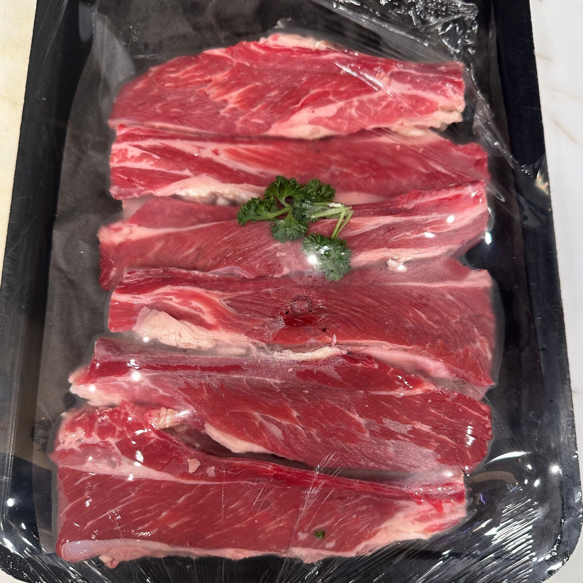 Raw beef short ribs with marbling, wrapped in clear plastic on a black tray with a small green parsley garnish in the center.