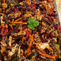 A close-up of a mixed vegetable stir-fry dish in a rectangular baking pan, featuring shredded cabbage, carrots, red peppers, and a small green broccoli garnish in a dark sauce.