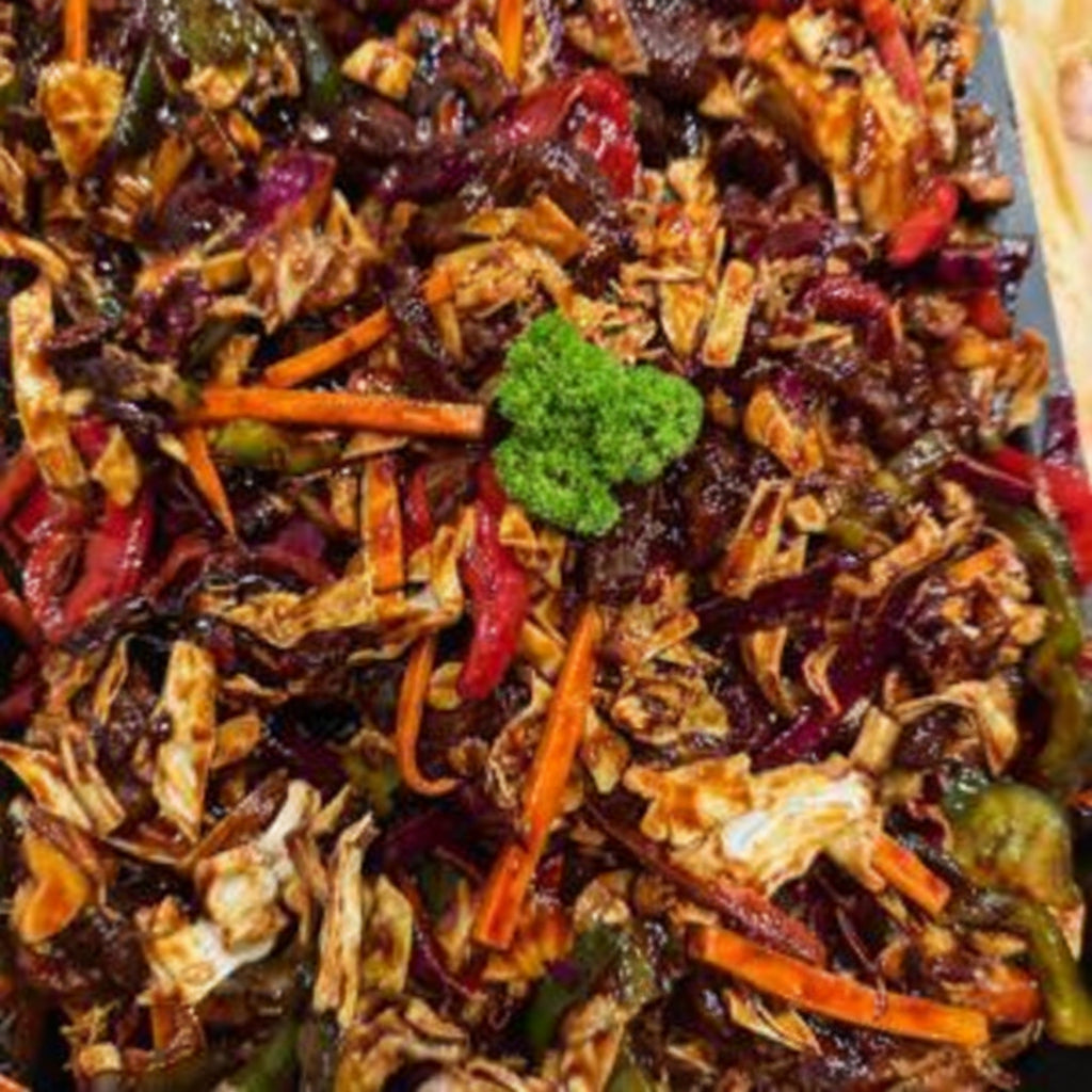 A close-up of a mixed vegetable stir-fry dish in a rectangular baking pan, featuring shredded cabbage, carrots, red peppers, and a small green broccoli garnish in a dark sauce.