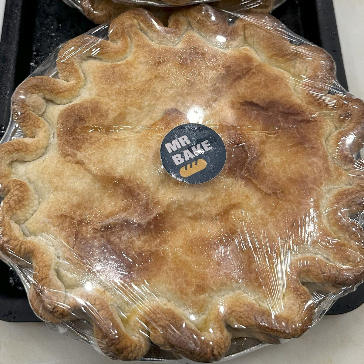 A baked pie wrapped in clear plastic with a round black and white 'MR BAKE' logo sticker in the center, sitting on a black tray.