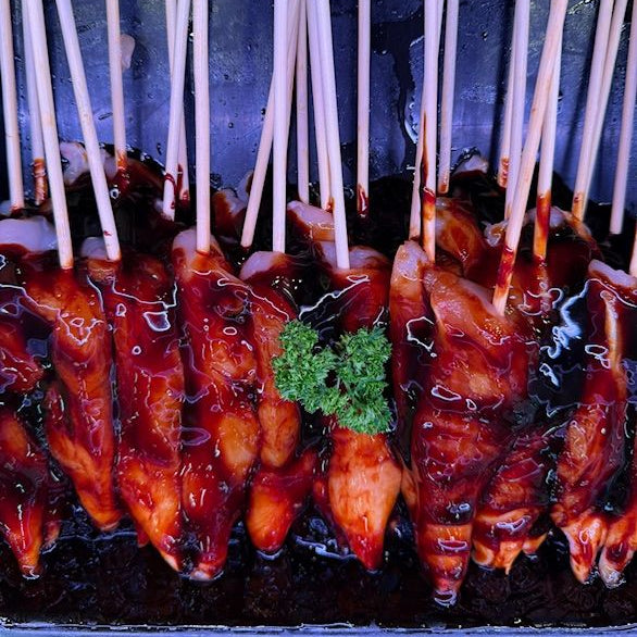 Close-up of several pieces of glazed chicken in honey soy on wooden skewers, arranged in a dark container with glossy sauce and a small green garnish in the center.