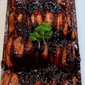 A close-up overhead shot of HONEY SOY NIBBLES coated in a glossy dark brown sauce and topped with a small green parsley garnish, presented in a clear plastic container.
