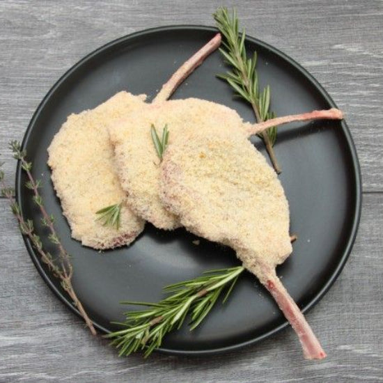 Three breaded lamb chops arranged on a round black plate with rosemary sprigs, photographed from above on a gray wood surface.