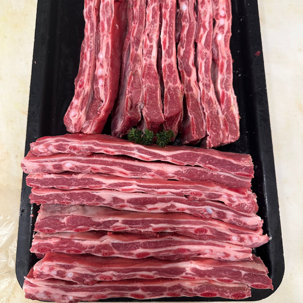 Raw lamb ribs arranged on a black tray, with vertical long strips on top and stacked horizontal strips below, photographed from above on a light beige surface.