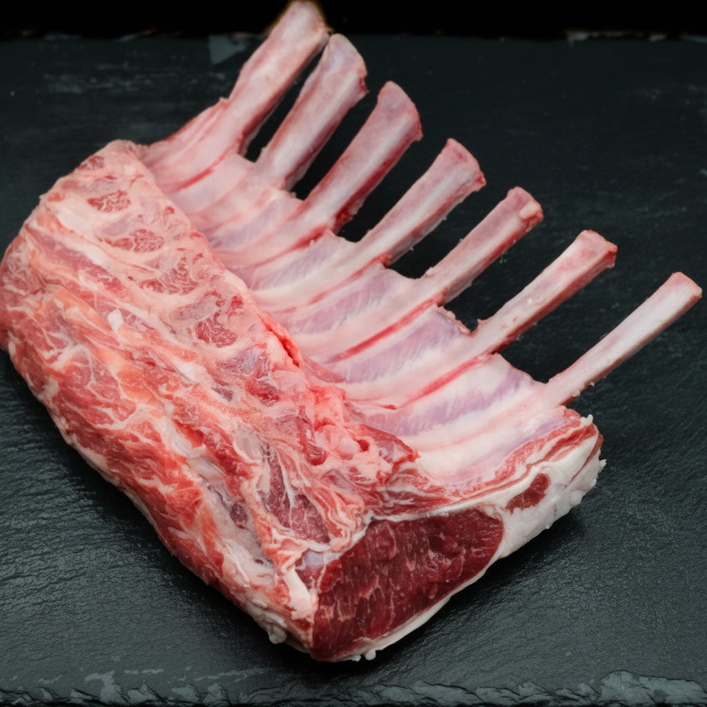 A raw rack of lamb with visible marbling and bone, photographed on a dark slate surface.