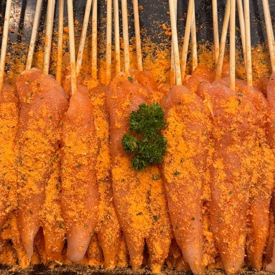 Raw chicken fillets on skewers, coated with macadamia nut and satay seasoning and sprinkled with a small green herb garnish, arranged on a dark tray.