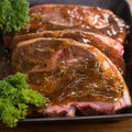 Four lamb chops marinated in a mint and rosemary sauce, garnished with fresh green parsley, presented in a black metal baking pan on a wooden surface.