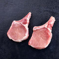 Two raw pork cutlets with bone, showing pink meat and white fat marbling, placed on a dark textured surface. Available at Robina Quality Meats