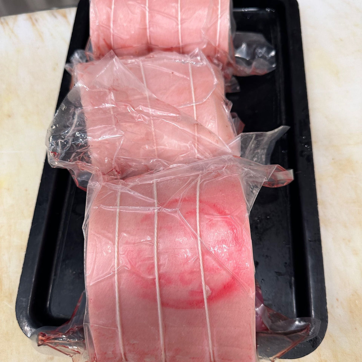 Three rolled pieces of pork loin roast, wrapped in clear plastic and tied with string, arranged on a black tray on a light beige countertop. Available to Robina Quality Meats