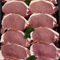 Multiple raw pork chops arranged in rows on a dark tray, with small green herb garnishes placed between some cuts.