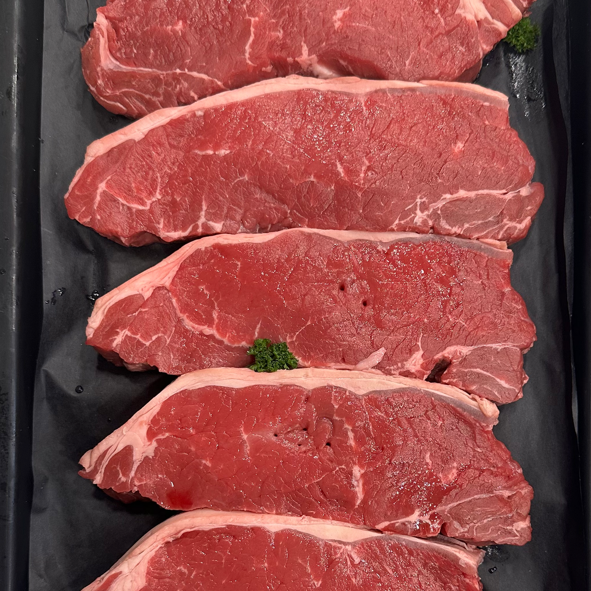 Six raw beef tenderloin steaks arranged vertically in a black plastic tray, garnished with small green parsley sprigs.