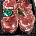 Four raw beef chuck steaks with visible fat marbling, sealed in a black plastic tray with a green 'Grass Fed' sticker, on a light beige surface.