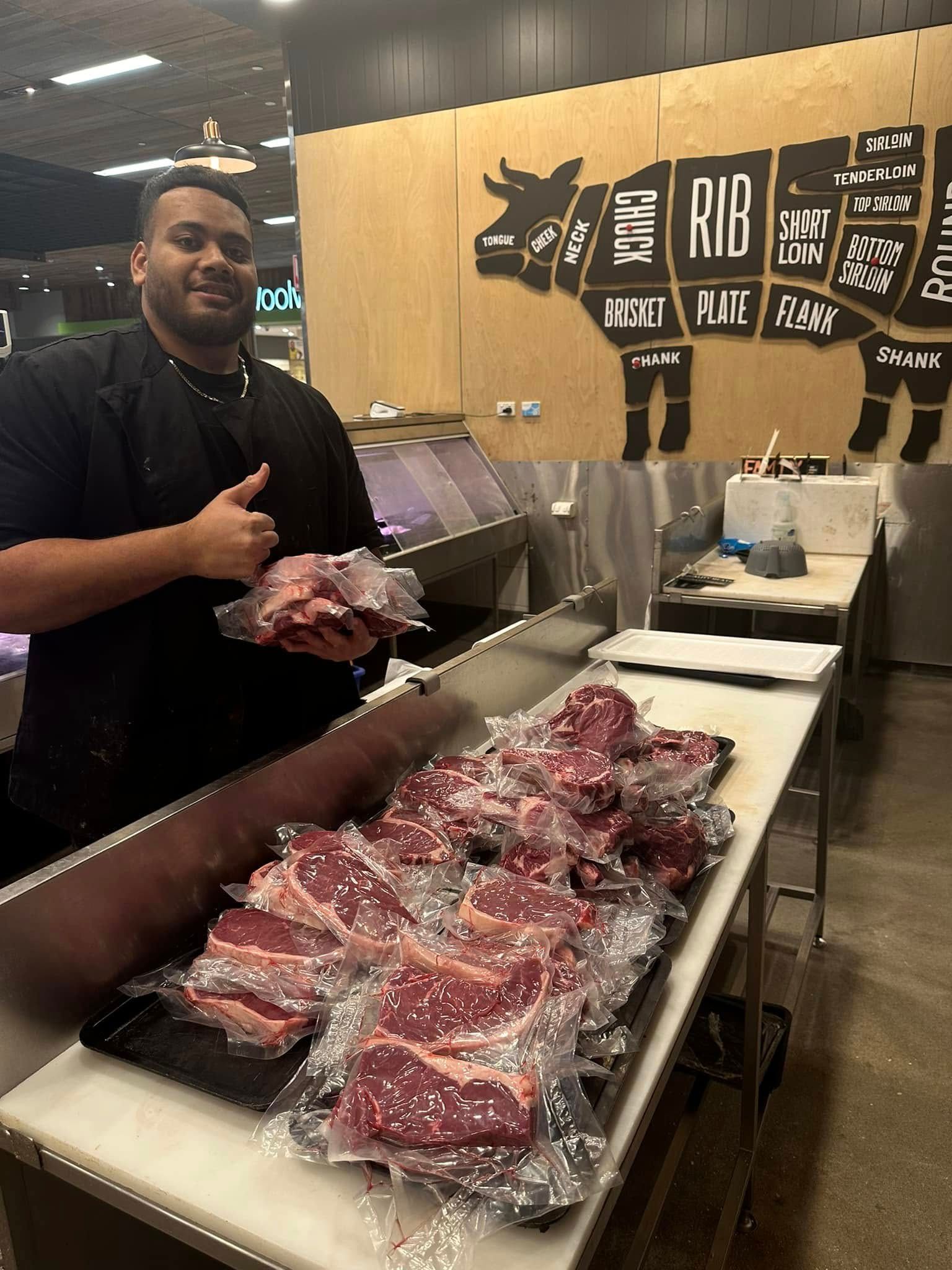 Gearge the lead butcher at Robina Quality Meats in a black chef's coat stands behind a butcher counter holding a vacuum-sealed package of raw meat, with multiple vacuum-sealed steaks displayed on the counter in a butcher shop. A large wooden wall sign behind him shows a cow diagram with labeled cuts.