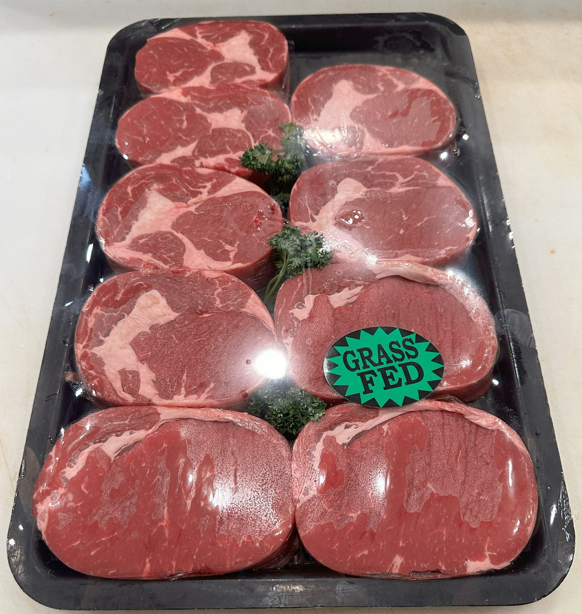 A black plastic tray holding eight raw beef steaks arranged in two rows, with a green circular 'GRASS FED' sticker on the front. The steaks have a deep red color with visible white fat marbling, and small green herb garnish is placed in the center.